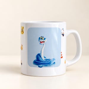 Może przedstawiać: Biały ceramiczny kubek z centralną prostokątną grafiką przedstawiającą niebieskiego węża z kreskówki z dużymi oczami i uśmiechniętymi ustami. Kubek posiada również mniejsze, rozproszone ilustracje różnych przedmiotów, w tym pachołka drogowego.