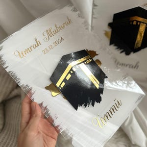 May include: A white acrylic plaque with gold lettering that reads "Umrah Mubarak 23.12.2024" and "Ummi". A black and gold cutout of the Kaaba is on the plaque.