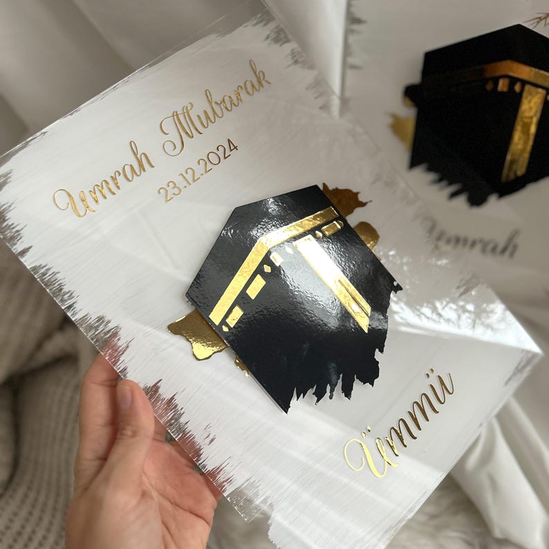 May include: A white acrylic plaque with gold lettering that reads "Umrah Mubarak 23.12.2024" and "Ummi". A black and gold cutout of the Kaaba is on the plaque.