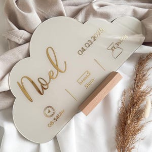 Könnte beinhalten: Ein weißes, wolkenförmiges Acrylschild mit goldener Beschriftung. Das Schild trägt die Aufschrift "Noel" und enthält das Datum "04.03.2025", die Uhrzeit "08:54Uhr" und die Maße "51cm".