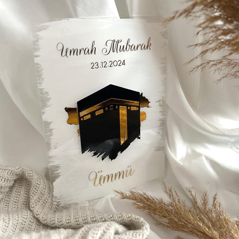 May include: White acrylic plaque with gold lettering and a black silhouette of the Kaaba. The text reads "Umrah Mubarak 23.12.2024 Ummi".