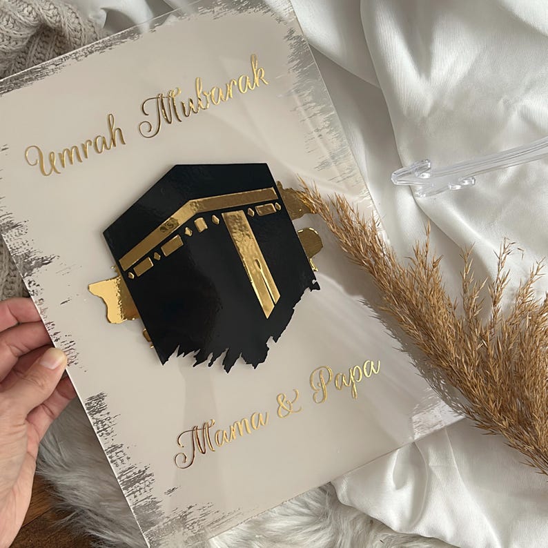 May include: A white acrylic plaque with gold lettering that reads "Umrah Mubarak" and "Mama & Papa". A black and gold 3D image of the Kaaba is in the center of the plaque.