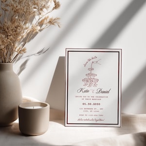May include: A rectangular wedding save the date card with a dark red border. The card features a line drawing of a champagne tower, the names Katie and Daniel, the date 01.30.2030, and text including an address and RSVP phone number.