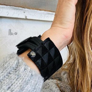 Könnte beinhalten: Ein schwarzes Armband mit Steppmuster und einem dekorativen Knopf. Das Armband besteht aus einem weichen, strukturierten Material und wird mit einer Schnalle befestigt. Das Bild enthält auch den handgeschriebenen Text "Ma-oo".