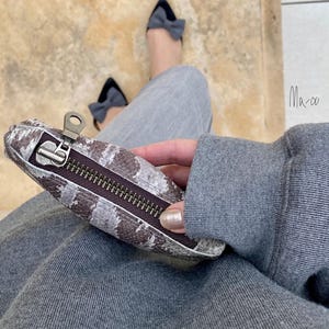 以下が含まれることがあります： グレーとブラウンのヘビ柄の小さなジッパー付きポーチで、シルバーのジッパーが付いています。ポーチは、グレーのパンツとグレーのセーターを着た人が持っています。