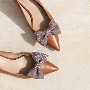 Può includere: Un paio di ballerine in pelle marrone con punta, ognuna ornata da un fiocco. I fiocchi sono realizzati in un materiale marrone e bianco a fantasia, aggiungendo un tocco decorativo alle scarpe. Le scarpe sono su uno sfondo neutro.