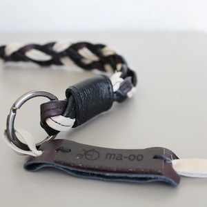 Op de afbeelding: Een leren sleutelhanger met een gevlochten band in zwart en wit. De sleutelhanger heeft een zilveren ring en een donkerbruin leren label met de tekst "ma-oo".