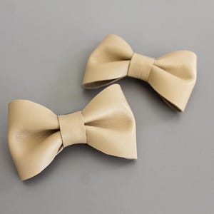 Può includere: Due fermagli per capelli in pelle beige a forma di fiocco. I fiocchi sono legati in un semplice nodo e hanno una finitura liscia e opaca.