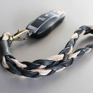Puede incluir: Un llavero de cuero trenzado negro y beige con un anillo plateado y un llavero de coche negro.