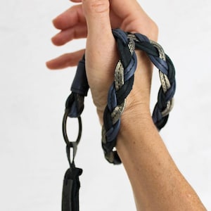 Op de afbeelding: Een gevlochten leren band met een zilveren en blauwe kleurstelling. De band heeft een metalen ring en een zwart leren tabblad.