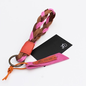 Op de afbeelding: Gevlochten leren sleutelhanger in bruin en roze met een oranje accent. De sleutelhanger heeft een metalen ring en een oranje leren lus. Een zwart label en een roze label met de tekst "ma-oo" zijn ook inbegrepen.