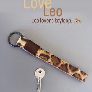 Può includere: Un portachiavi in ​​cuoio marrone con un design a stampa leopardata. Il portachiavi ha un anello di metallo e il testo "Love Leo" e "Leo lovers keyloop..." è visibile sopra il portachiavi.