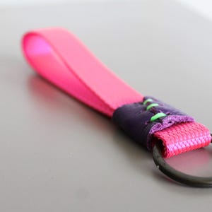 May include: A bright pink keychain wrist strap with a dark purple leather accent and a black metal ring. The strap is made of a woven material and is stitched with green thread.