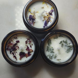 May include: Three small glass jars with lids, each containing a white candle with different dried flower petals. The jars are arranged in a triangle shape.