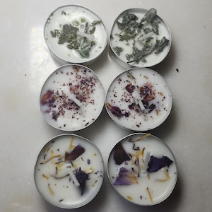 May include: Six small, round candles in silver metal containers. The white wax candles are decorated with dried herbs and flower petals. The candles are arranged in two rows of three.