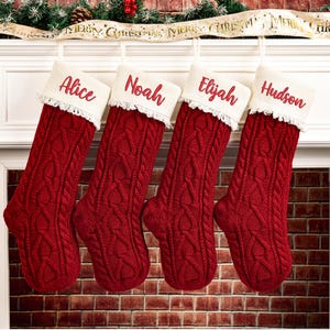Puede incluir: Cuatro calcetines navideños rojos de punto trenzado cuelgan de una repisa blanca. Cada calcetín tiene un puño blanco con ribete de encaje y un nombre bordado en rojo: Alice, Noah, Elijah y Hudson. Una cinta festiva con "Merry Christmas" está arriba.
