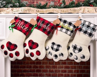 May include: Four Christmas stockings shaped like paw prints, each with a name embroidered on the cuff. The stockings have a white, fluffy body and a plaid cuff in red and black or black and white. Each stocking has a heart-shaped paw pad.
