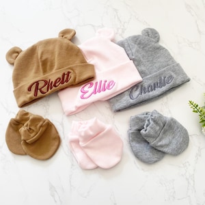 May include: Three personalized baby hats in brown, pink, and gray with the names Rhett, Ellie, and Charlie embroidered on them.  The hats have bear ears and come with matching mittens.