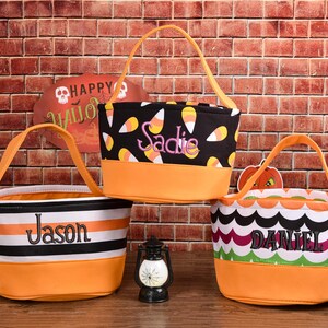 May include: Three Halloween-themed treat bags with orange handles and bases. One bag is black with candy corn, another has black and white stripes, and the third has a scalloped pattern. Each bag has a name embroidered on it.