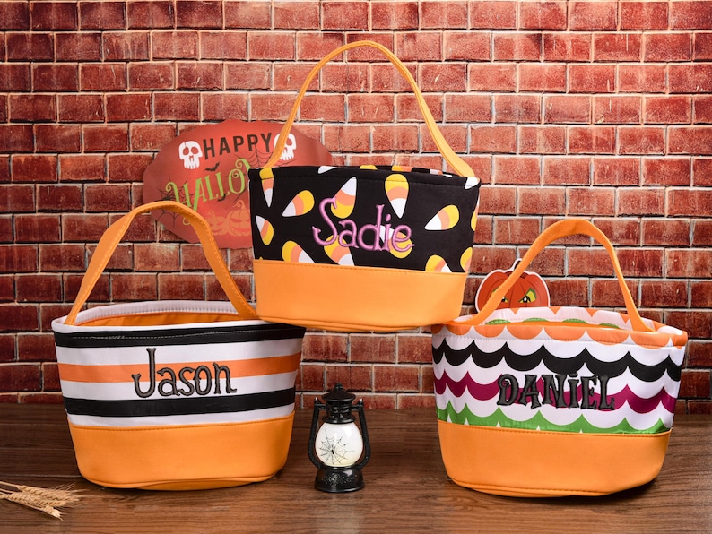 May include: Three Halloween-themed treat bags with orange handles and bases. One bag is black with candy corn, another has black and white stripes, and the third has a scalloped pattern. Each bag has a name embroidered on it.