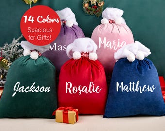 May include: A collection of colorful velvet Santa sacks in various shades, including green, purple, red, pink, and blue. Each sack is personalized with a name in white script and topped with a white faux fur trim and pom-poms. The image includes text that says "14 Colors Spacious for Gifts!"