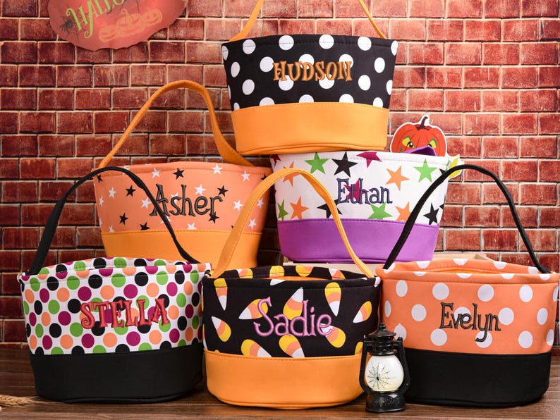 May include: A collection of personalized Halloween treat bags. Each bag features a unique design, including polka dots, stars, and candy corn patterns, with names embroidered on them. The bags have orange, black, and purple accents and handles.
