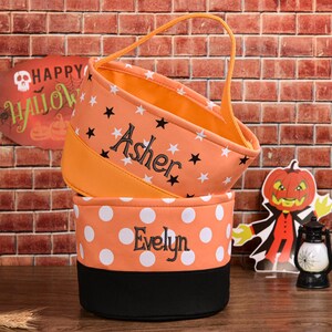 May include: Two orange Halloween treat bags, one with stars and the name "Asher", the other with white polka dots and the name "Evelyn". A handle is attached to the top bag. A sign that says "Happy Halloween" is in the background.