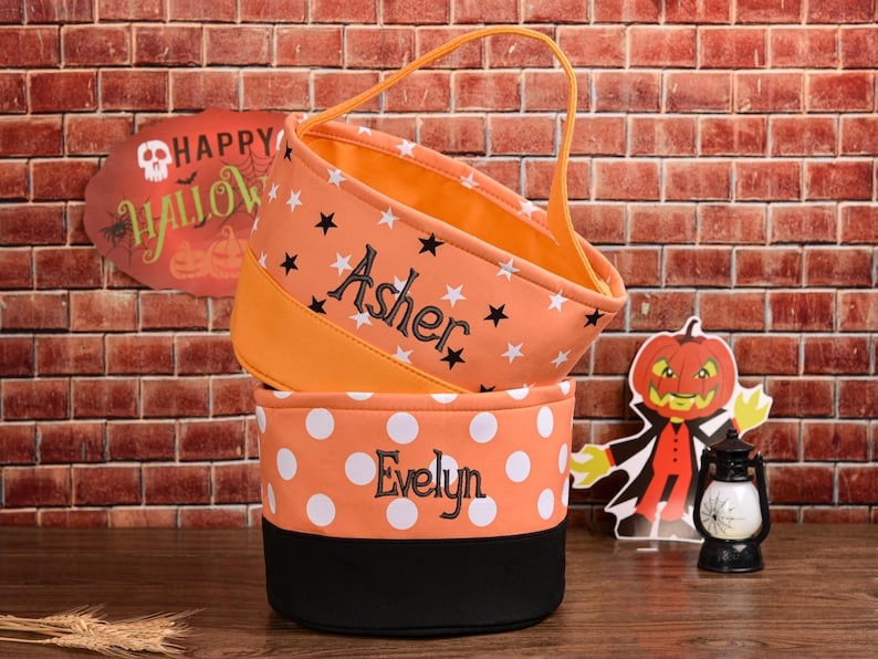 May include: Two orange Halloween treat bags, one with stars and the name "Asher", the other with white polka dots and the name "Evelyn". A handle is attached to the top bag. A sign that says "Happy Halloween" is in the background.