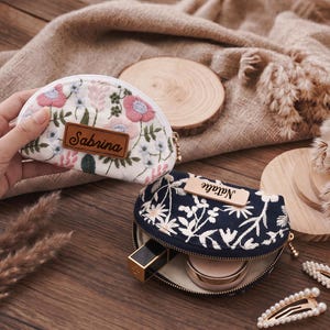 May include: Two semi-circular zippered pouches featuring floral embroidery. One pouch is white with pink and blue flowers and a brown leather tag reading Sabrina. The other is navy blue with white floral patterns and a light wood tag reading Natalie.