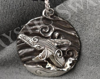 May include: A silver pendant necklace with a round charm featuring a whale jumping out of the water. The charm has a textured surface and is detailed with swirling waves.
