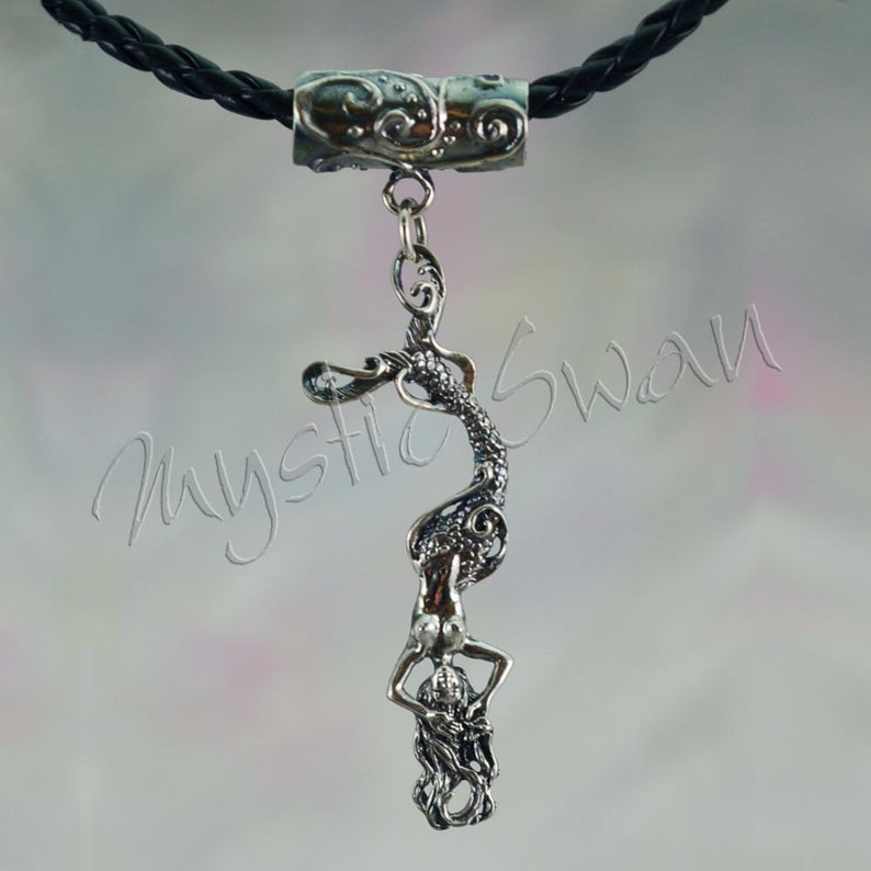 May include: Silver pendant necklace featuring a detailed mermaid design. The pendant is suspended from a black braided cord. The mermaid is depicted with flowing hair and a detailed tail. The necklace has a decorative silver clasp.