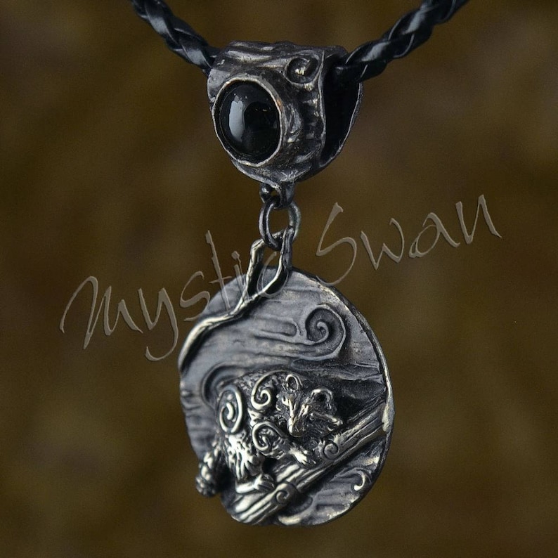Puede incluir: Un collar de plata con un colgante de &oacute;nix negro y un dije de plata detallado de un mapache en una rama. El dije est&aacute; colocado sobre un fondo texturizado con dise&ntilde;os en espiral.