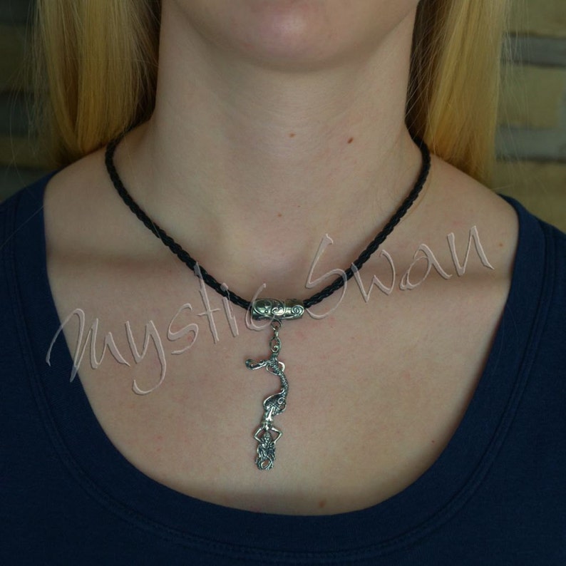 May include: A black braided cord necklace with a silver pendant. The pendant features a stylized figure of a woman. The necklace is worn against a dark blue shirt.