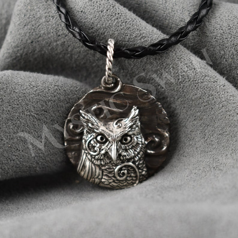 May include: A silver owl pendant with intricate detailing, suspended from a black braided leather cord. The owl is centered on a circular, textured background. The pendant's design features detailed feathers and large, expressive eyes.