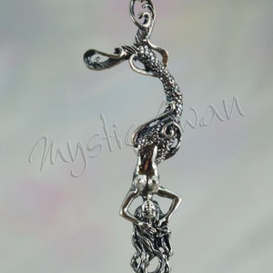 May include: A silver-toned pendant featuring a mermaid design. The mermaid is depicted upside down, with her tail curving upwards and her body extending downwards. The pendant has intricate detailing, including scales and flowing hair.