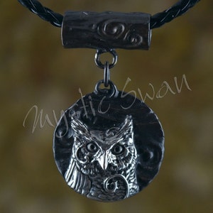 May include: A silver-toned pendant necklace featuring an owl design. The pendant is circular with a detailed owl face in the center. The necklace has a black braided cord and a decorative cylindrical clasp. The text "mystic swan" is visible.