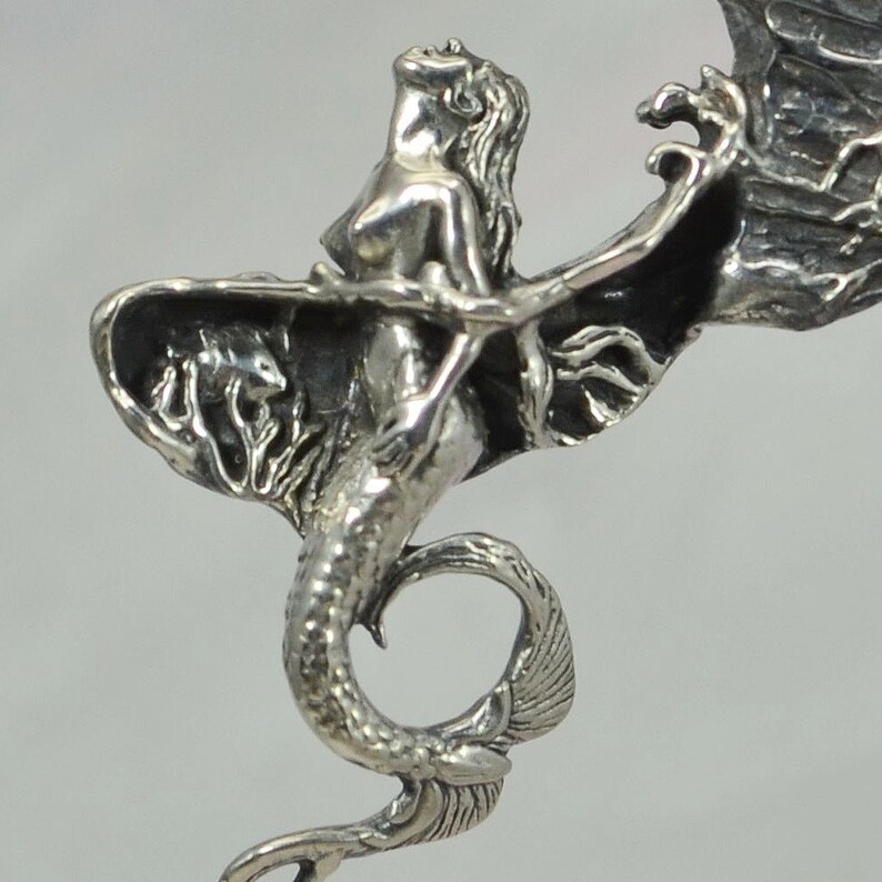 May include: A silver pendant featuring a mermaid with wings, her tail curled around her body. The mermaid is depicted with a flowing tail and a delicate, detailed design.