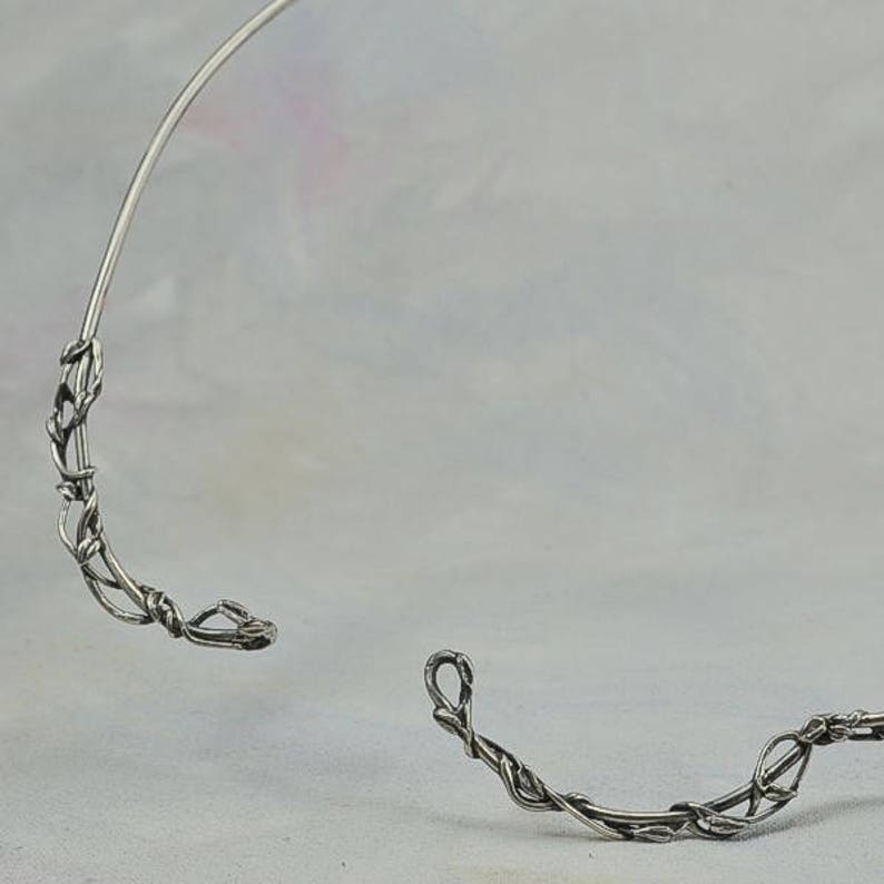 May include: A silver wire choker necklace with a delicate, vine-like design. The necklace is open in the back and has a simple, elegant style.