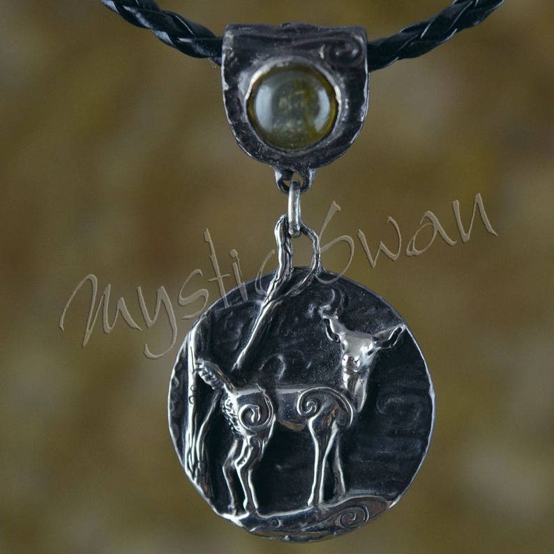Puede incluir: Un collar con un colgante de plata en un cord&oacute;n de cuero negro. El colgante presenta una representaci&oacute;n detallada de un ciervo con una piedra preciosa verde en el centro de la parte superior del colgante.