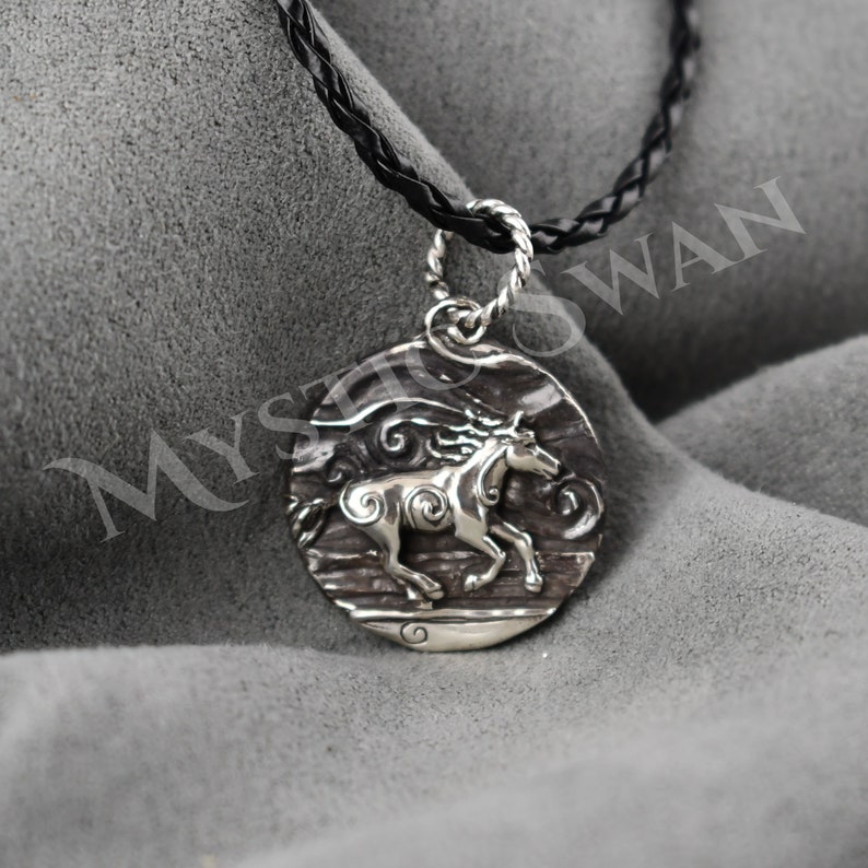 Puede incluir: Collar con colgante de plata con un dise&ntilde;o detallado de un caballo corriendo a trav&eacute;s de olas arremolinadas. El collar est&aacute; en un cord&oacute;n trenzado negro.