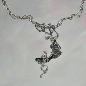 May include: A silver necklace with a detailed pendant featuring a mermaid and a figure climbing a rocky cliff. The necklace is made of twisted silver wire.