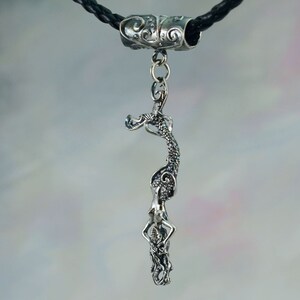 May include: A silver mermaid pendant necklace with a black braided leather cord. The pendant features a detailed mermaid design with intricate scales and flowing hair. The necklace has a decorative silver clasp.