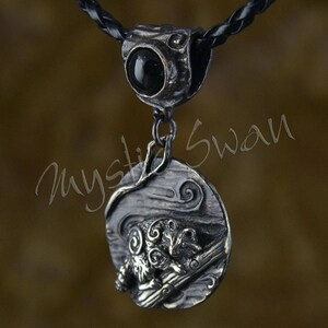May include: A silver-toned pendant necklace with a black stone and a detailed ram design. The pendant hangs from a black braided cord. The ram is depicted in a detailed, artistic style, with swirling patterns and a textured background.