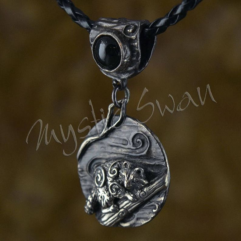 May include: A silver-toned pendant necklace with a black stone and a detailed ram design. The pendant hangs from a black braided cord. The ram is depicted in a detailed, artistic style, with swirling patterns and a textured background.