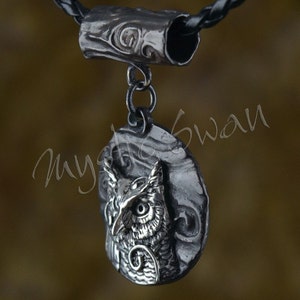 May include: A silver-toned owl pendant with intricate detailing, suspended from a black leather cord. The owl is set against a textured background, and the pendant is connected to a decorative cylindrical bail.