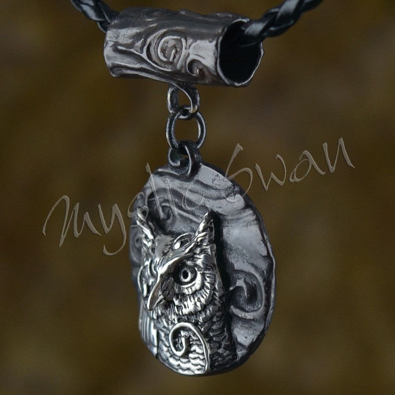 May include: A silver-toned owl pendant with intricate detailing, suspended from a black leather cord. The owl is set against a textured background, and the pendant is connected to a decorative cylindrical bail.