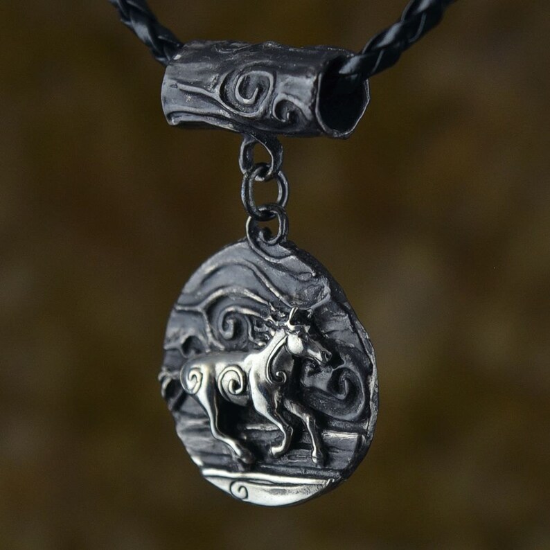Puede incluir: Un collar con un colgante de plata de dise&ntilde;o circular que presenta un caballo estilizado corriendo a trav&eacute;s de l&iacute;neas en espiral. El colgante est&aacute; unido a un cord&oacute;n de cuero negro con un conector de barril de plata.