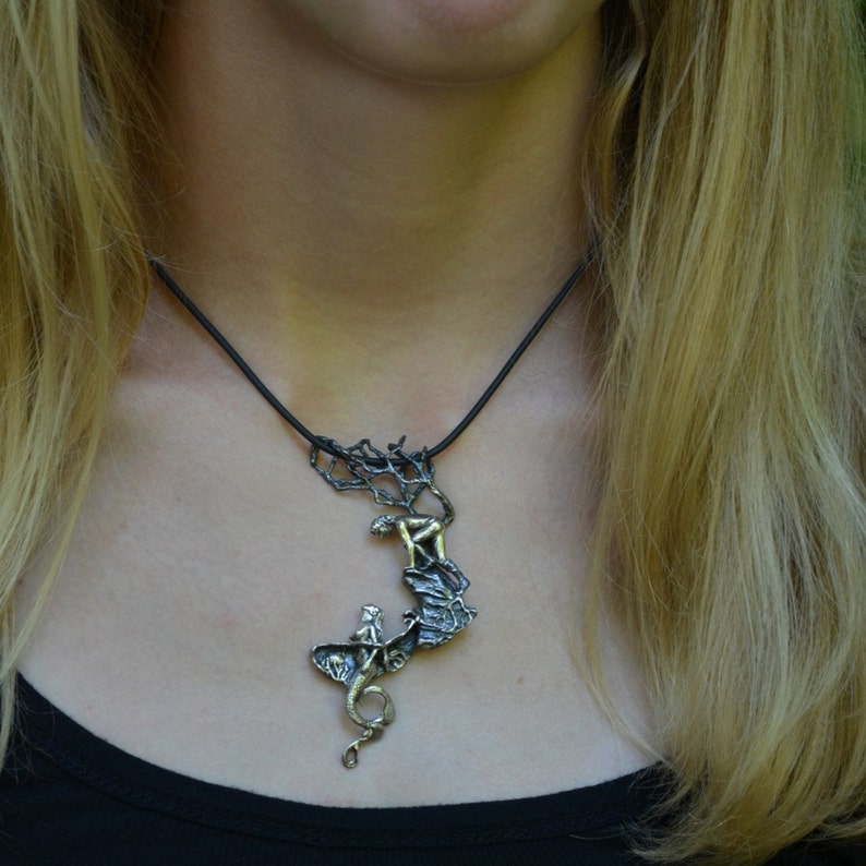 May include: A silver necklace with a pendant featuring a woman climbing a tree with a dragon at the bottom. The pendant is made of metal and has a detailed design.