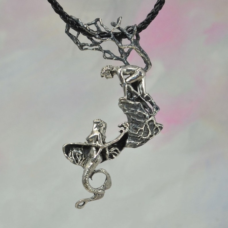 May include: A silver pendant necklace featuring a sculpted mermaid with a human-like torso and a fish tail, and a man climbing a rock formation. The pendant is attached to a black cord.