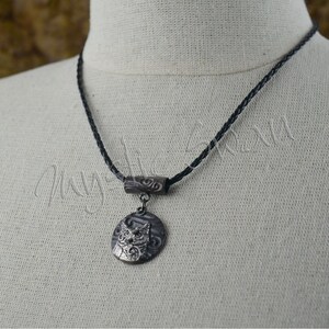 May include: A silver-toned pendant necklace featuring an owl design. The pendant hangs from a black braided cord. The owl is detailed with etched features and is set against a textured background. The necklace has a decorative bar above the pendant.